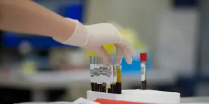 Gloved hand picking up blood sample test tubes in a laboratory with blurred background of lab equipment.