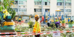 Construction workers clearing debris in an urban playground area near apartment buildings under bright daylight.