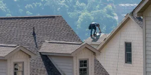 Roofer working on shingled roof of a beige house surrounded by green forested hills in the background.