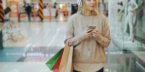 Young woman with headphones holding phone and shopping bags walking inside a bright mall corridor.
