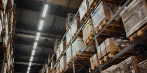 Rows of tall shelves with stacked pallets of boxed goods in a large industrial warehouse under bright lights