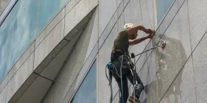 Worker wearing safety gear cleaning windows on a tall modern building exterior using ropes and harness.