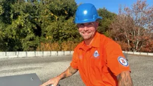 Smiling construction worker wearing blue hard hat and orange shirt working outdoors with tools on rooftop.