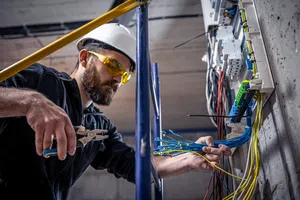 Electrician wearing safety gear working with blue and yellow electrical wires in an industrial setting.