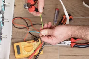 Electrician using wire cutters to strip electrical wires beside a yellow multimeter on wooden floor