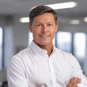 Professional man wearing a white shirt smiling confidently with arms crossed in a blurred modern office background.