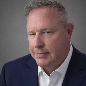 Middle-aged man in a navy blue suit and white shirt looking confidently at the camera against a gray background