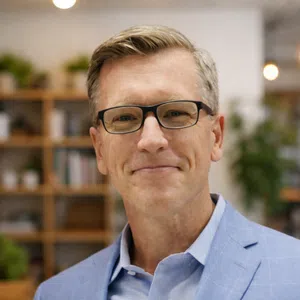 Middle-aged man wearing glasses and a blue blazer smiling in a modern office with shelves and plants.