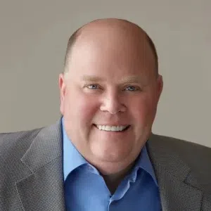 Smiling middle-aged bald man wearing a blue shirt and gray blazer against a neutral background