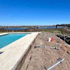 Poolside construction site with metal anchors, a red excavator, and a waterfront view under a clear blue sky