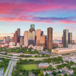 Aerial view of Houston skyline with highways and green spaces during a colorful sunset sky.