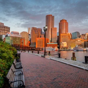 Boston waterfront skyline at sunset with benches, brick walkway, and riverfront boats visible.