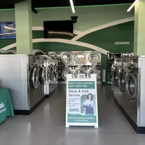 Modern laundromat interior with rows of stainless steel washers and dryers and wash & fold service sign.