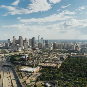 Aerial view of a city skyline with highways, residential areas, green spaces, and a partly cloudy blue sky.