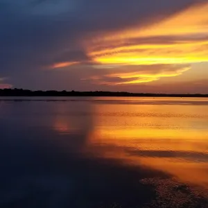 Calm lake at sunset with orange and yellow skies reflecting on still water under dark clouds.