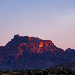 Sunset illuminates a rugged mountain towering over a small town in the desert landscape under a clear sky.