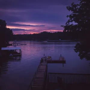 Tranquil lake at dusk with purple skies, silhouette trees, and wooden docks extending over calm water.