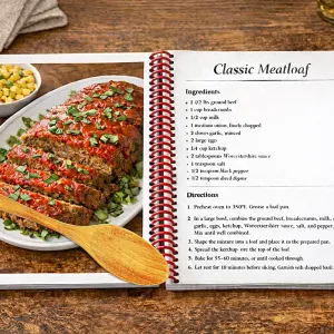 Open cookbook showing classic meatloaf recipe with sliced meatloaf and bowl of corn on wooden table.