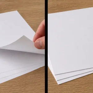 A hand flipping through a stack of text weight paper next to a stack of heavier cover paper on a wooden surface