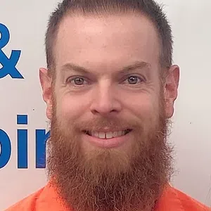 Smiling man with a red beard and styled hair wearing an orange shirt against a white background with blue text.