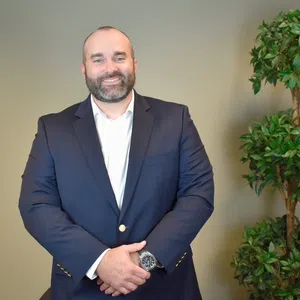 Man in a navy blue suit and white shirt smiling with hands clasped, standing beside a green indoor plant.