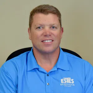 Man wearing a blue Estes Services polo shirt smiling against a plain beige background.
