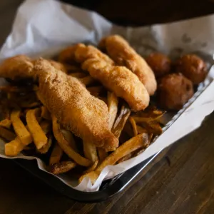 Basket of crispy fried fish, golden French fries, and hush puppies served on a tray with parchment paper.