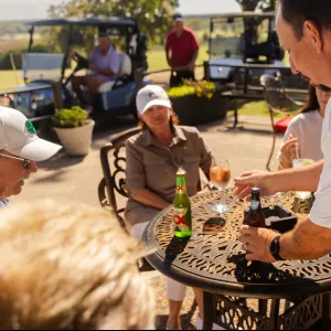 Group of people enjoying drinks and socializing outdoors at a golf course patio with golf carts in background