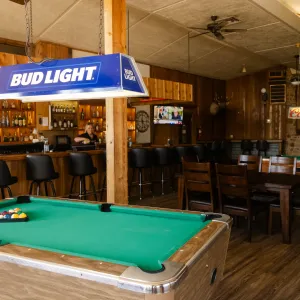 Cozy bar interior with pool tables, Bud Light and Coors Light signs, wooden furniture, and a woman sitting at the bar.
