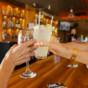 Two women clinking glasses with cocktails at a wooden bar in a cozy pub setting.