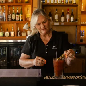 Bartender preparing a Bloody Mary cocktail with olives at a bar with liquor bottles in the background