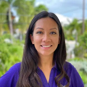 Smiling woman with long dark hair wearing a purple polo shirt outdoors with greenery and sky in background.