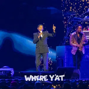 Singer performing on stage with saxophonist and DJ, vibrant lighting and audience visible in foreground