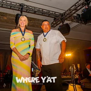 Two award recipients wearing medals stand on stage at a formal event with audience in background.