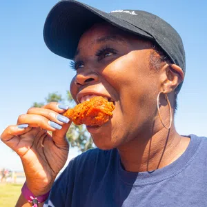 Smiling woman wearing a black cap and hoop earrings eating fried chicken outdoors on a sunny day
