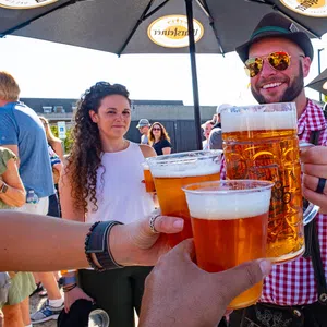 Group of people clinking large beer mugs and cups at an outdoor festival under branded umbrellas