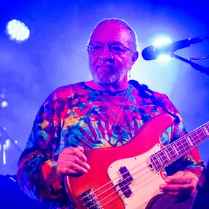 Musician wearing a colorful tie-dye shirt playing red electric bass guitar on stage with blue lighting.