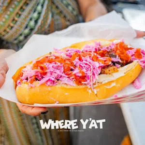 Close-up of a hand holding a hot dog with pink slaw and red sauce in a paper tray, with overlaid text reading WHERE Y'AT.