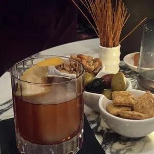 Old fashioned cocktail with lemon peel served with assorted nuts, olives, crackers, and breadsticks on marble table.
