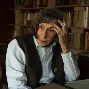 Elderly woman with brown hair sitting contemplatively in a vintage study room filled with books and a typewriter.