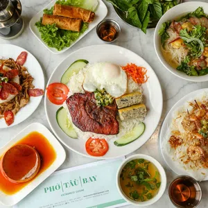 Overhead view of Vietnamese dishes including grilled pork with egg, spring rolls, noodle soup, shrimp skewers, and caramel flan on a marble table.