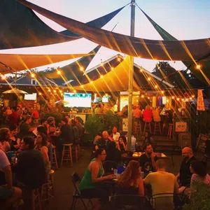 Outdoor evening bar scene with string lights, shaded canopies, and groups of people socializing under warm lighting.