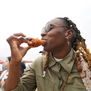 Person with braided hair and sunglasses enjoying crispy fried chicken outdoors at a casual event.