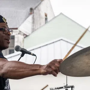 Man wearing a beret playing drums energetically at an outdoor performance with houses in the background