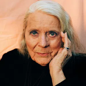 Elderly woman with silver hair, wearing black, resting her face on her hand against a soft peach background.