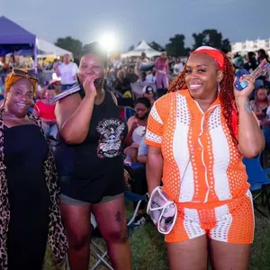 Three smiling women at an outdoor event with tents and crowd in the background during evening.