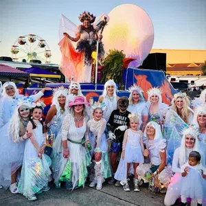 Group of people in white costumes and wings posing outdoors at a festival with a ferris wheel and colorful float.