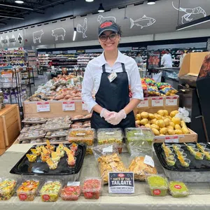 Smiling store employee presenting various homemade dips and salsas at a grocery store sampling table.