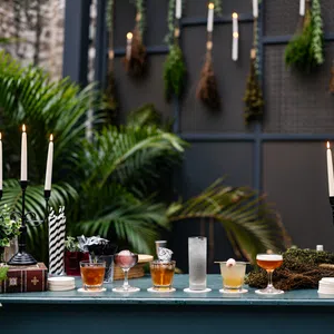 Outdoor bar setup with assorted cocktails, lit candles, greenery, and dried herbs hanging on a dark backdrop.