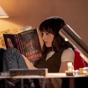 Woman reading the book Hospitality by Verity Crawford in a cozy, warmly lit room with a table lamp.